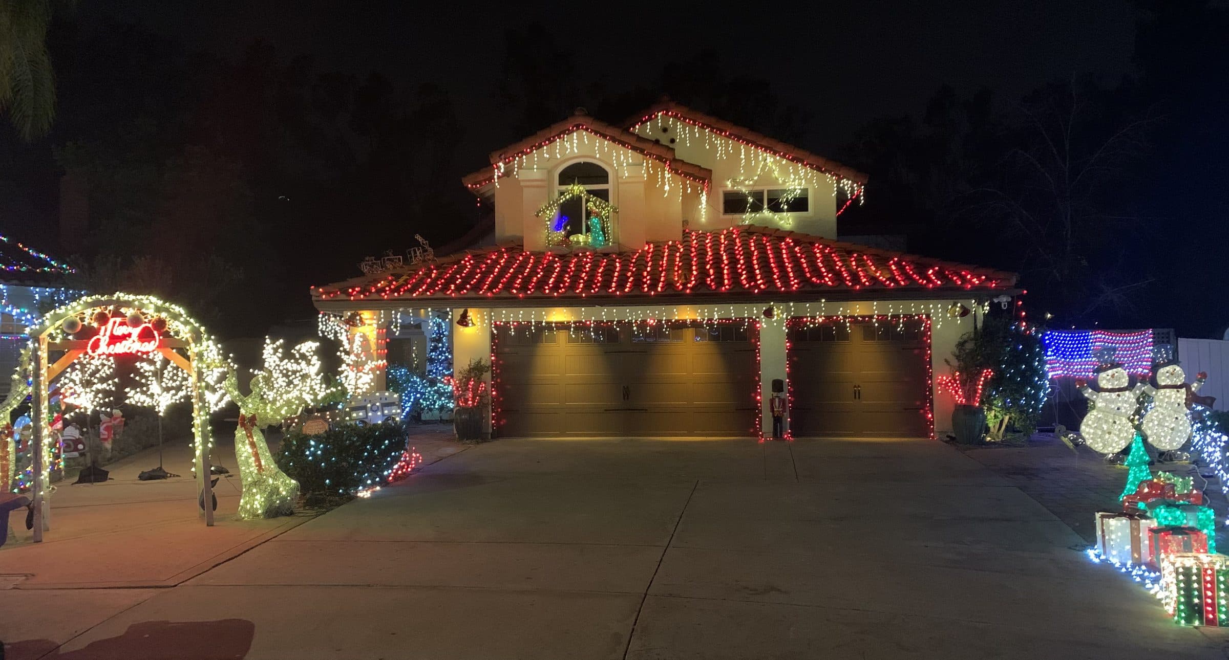 Justin's family's two-story house at night: red bulbs on every row of the tile roof, warm white icicle lights along the eaves and garage, a lit arch, reindeer, snowmen and gift boxes in the yard.