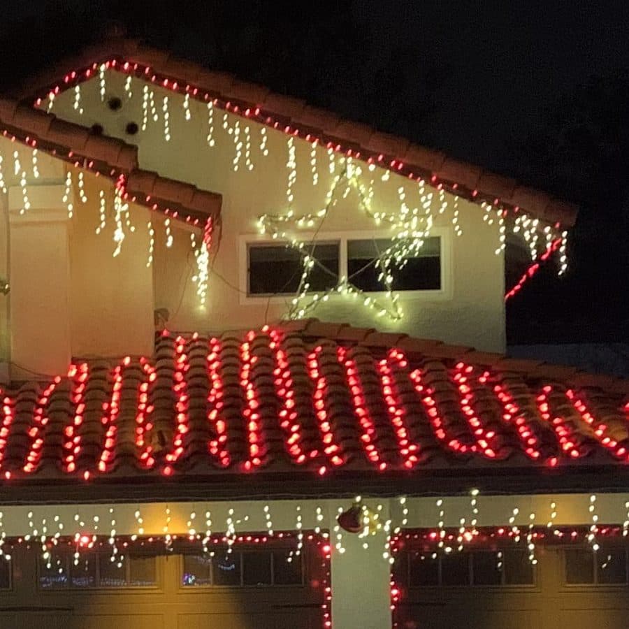 Close up of red C9 bulbs clipped along each row of a red tile roof, with warm white icicle lights on the eaves above.